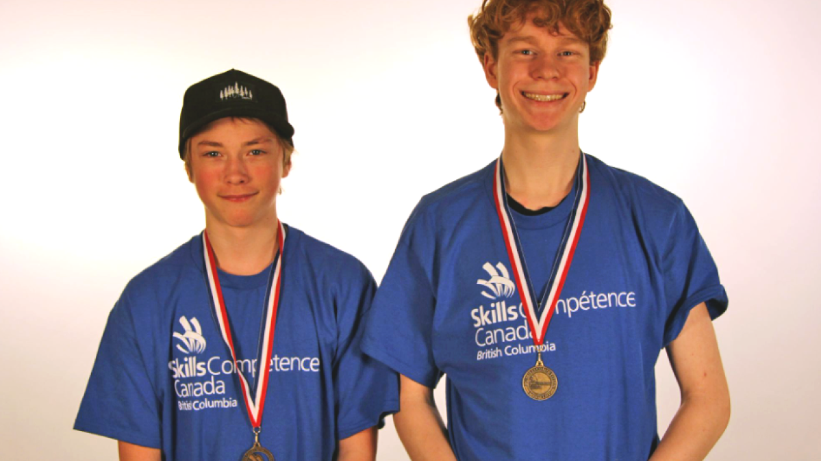 Two teenaged boys in blue t-shirts wearing gold medals and standing in front of a white background.