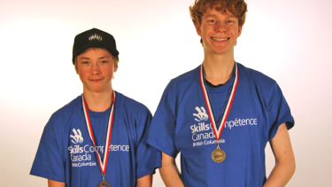 Two teenaged boys in blue t-shirts wearing gold medals and standing in front of a white background.
